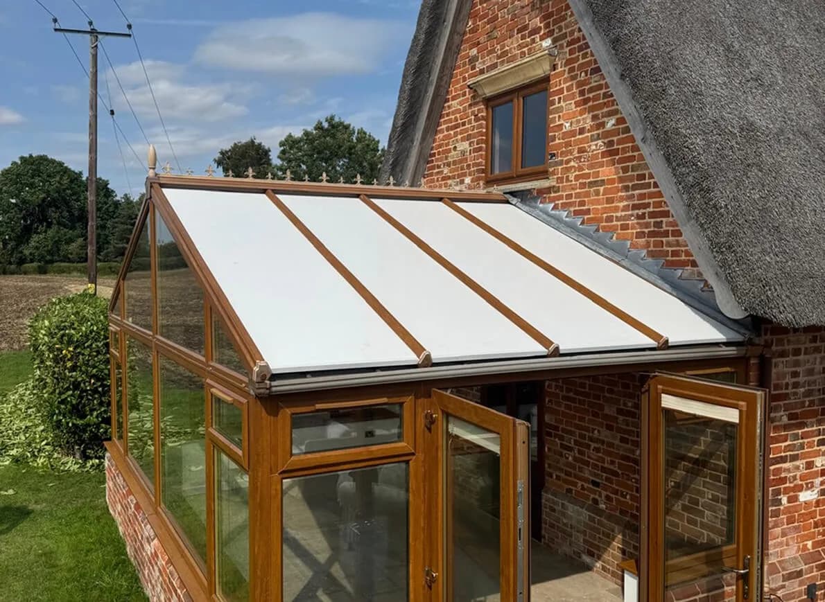 An insulated panel conservatory roof in white composite panels, between golden oak glazing bars, on a lean-to conservatory against a brick and thatch cottage.